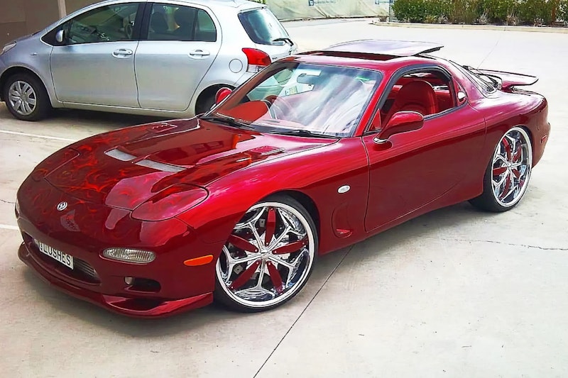 Front three-quarter view of a red Mazda RX-7 with flame graphics on the bonnet and chrome wheels with red inserts, parked outdoors next to a silver hatchback.