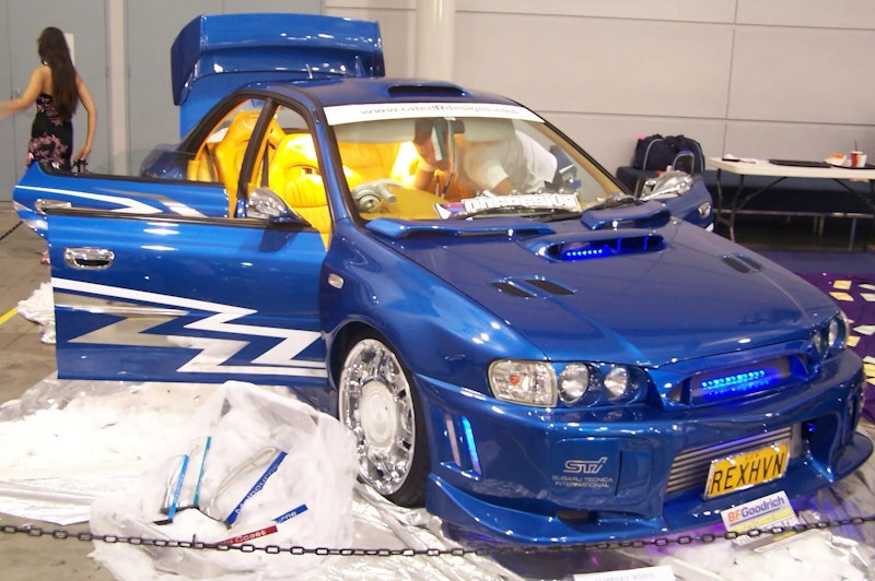 Front three-quarter view of a blue Subaru Impreza WRX STI with custom yellow lightning bolt graphics on the door, yellow leather interior, and "REXHVN" number plate, displayed on a cotton-cloud stand at an indoor car show.