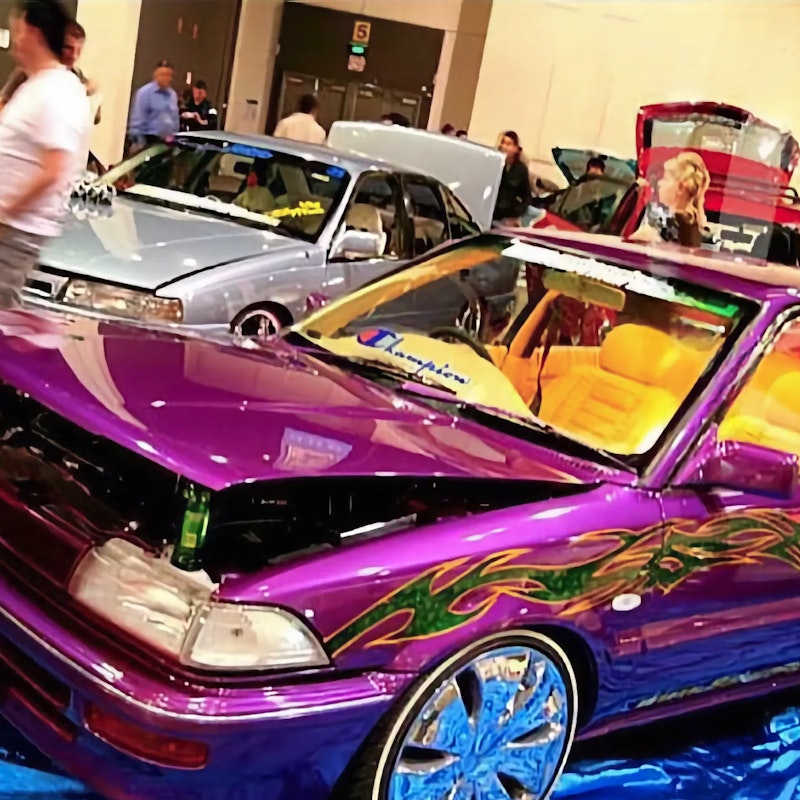 Purple show car with green flame and tribal graphics, hood up and custom rims, displayed on a blue tarp at an indoor car show with onlookers and other modified cars in the background.