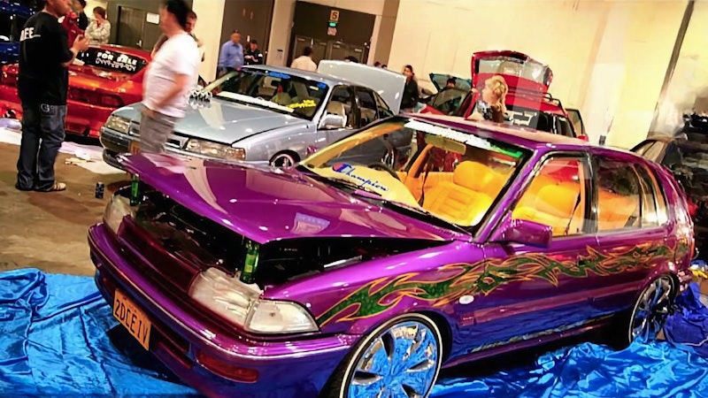 Purple show car with green flame and tribal graphics, hood up and custom rims, displayed on a blue tarp at an indoor car show with onlookers and other modified cars in the background.