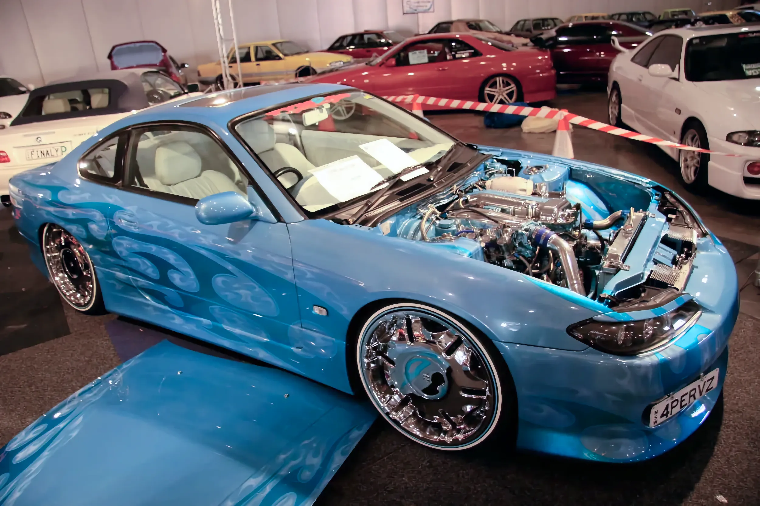 Light blue Nissan Silvia S15 with ghost flame graphics, white leather interior, and large chrome wheels, displayed with its hood open at an indoor car show surrounded by other parked cars.
