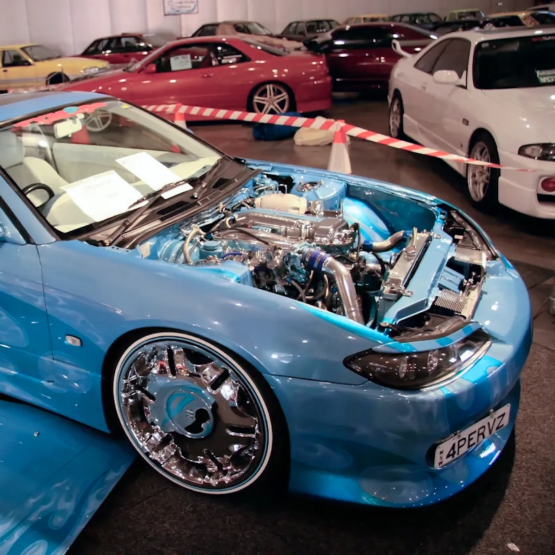Light blue Nissan Silvia S15 with ghost flame graphics, white leather interior, and large chrome wheels, displayed with its hood open at an indoor car show surrounded by other parked cars.
