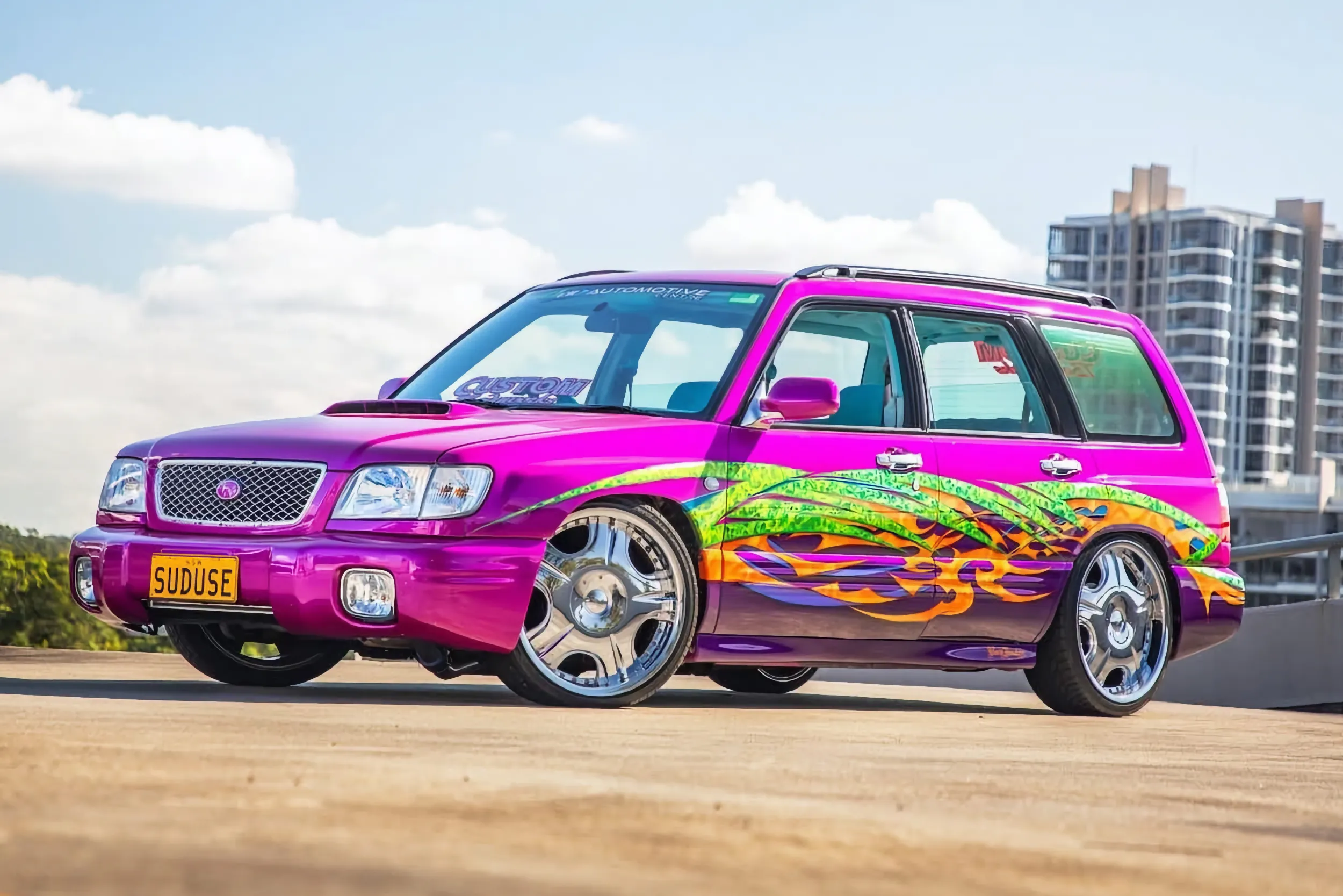 Pink and purple Subaru Forester with colourful green, orange, and yellow tribal flame graphics and chrome wheels, lowered and parked on a rooftop car park against a blue sky with an apartment tower in the background.