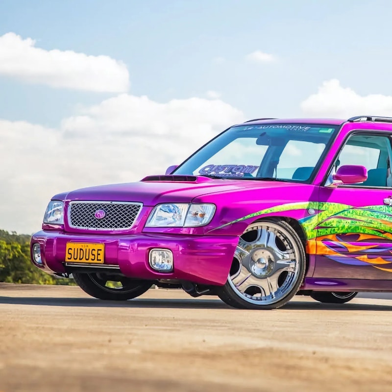 Pink and purple Subaru Forester with colourful green, orange, and yellow tribal flame graphics and chrome wheels, lowered and parked on a rooftop car park against a blue sky with an apartment tower in the background.