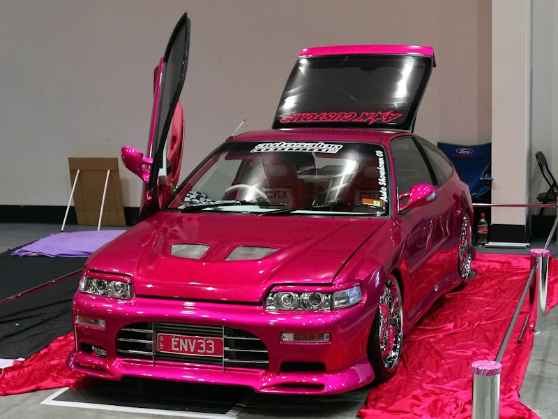 Bright pink Honda CRX with a vertical lambo-style door raised, chrome wheels, and "AK Customs" and "autosalon" windscreen banners (plate "ENV33"), displayed on red fabric at an indoor car show.