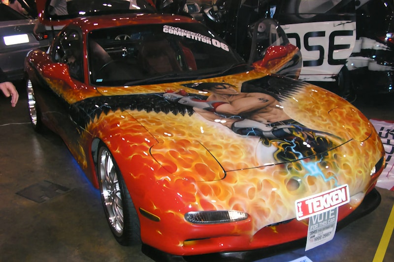 Red Mazda RX-7 with an elaborate airbrushed flame paint job and a Tekken video game character mural across the bonnet, "TEKKEN" front plate and a "Vote For This Car" sign, displayed at an indoor car show.