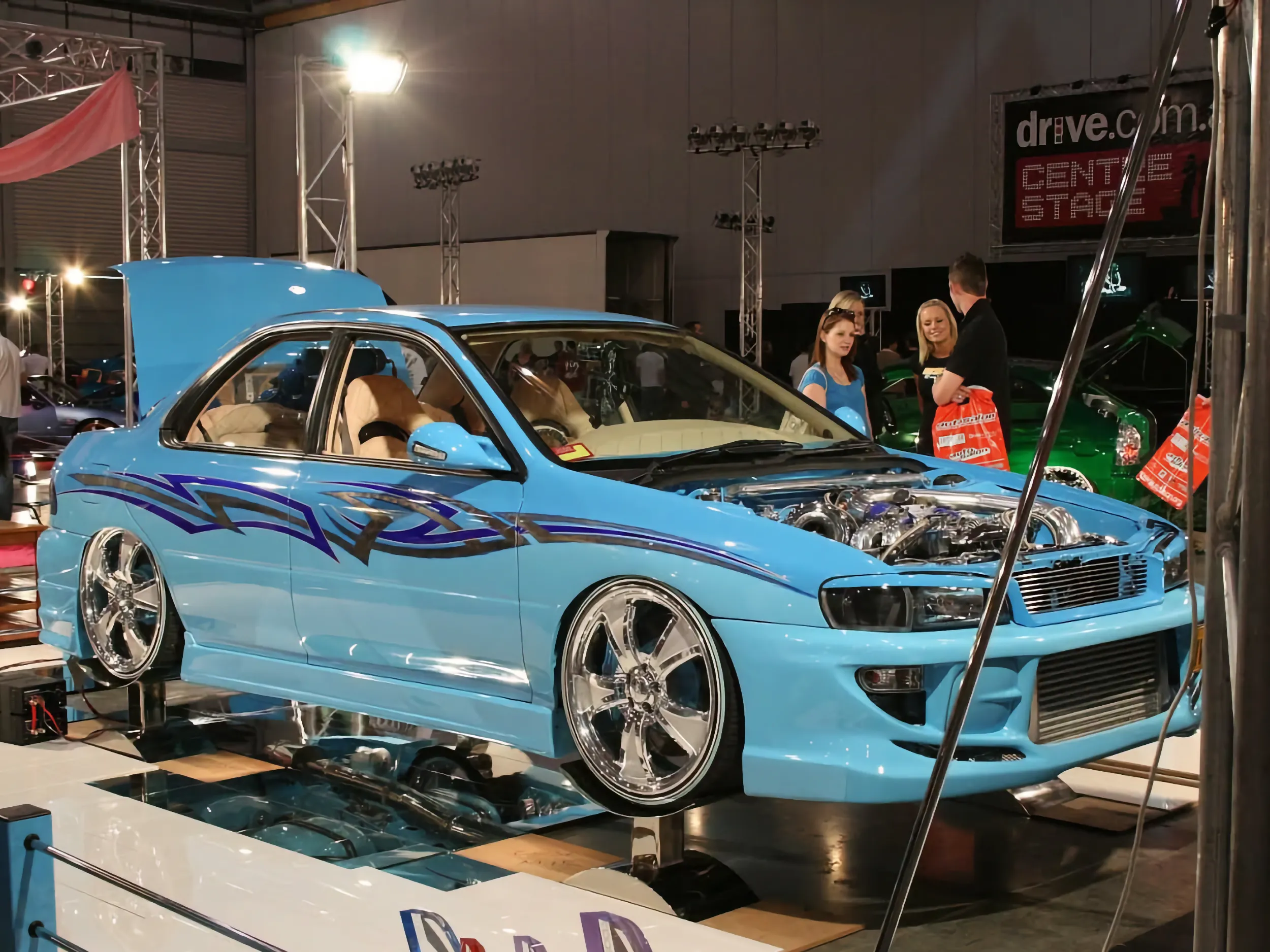 Blue custom Subaru Impreza with blue tribal graphics displayed on a mirrored plinth with its bonnet and doors open at an indoor car show, showing its chrome turbo engine and tan interior.
