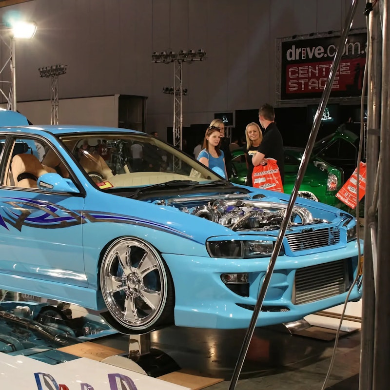 Blue custom Subaru Impreza with blue tribal graphics displayed on a mirrored plinth with its bonnet and doors open at an indoor car show, showing its chrome turbo engine and tan interior.