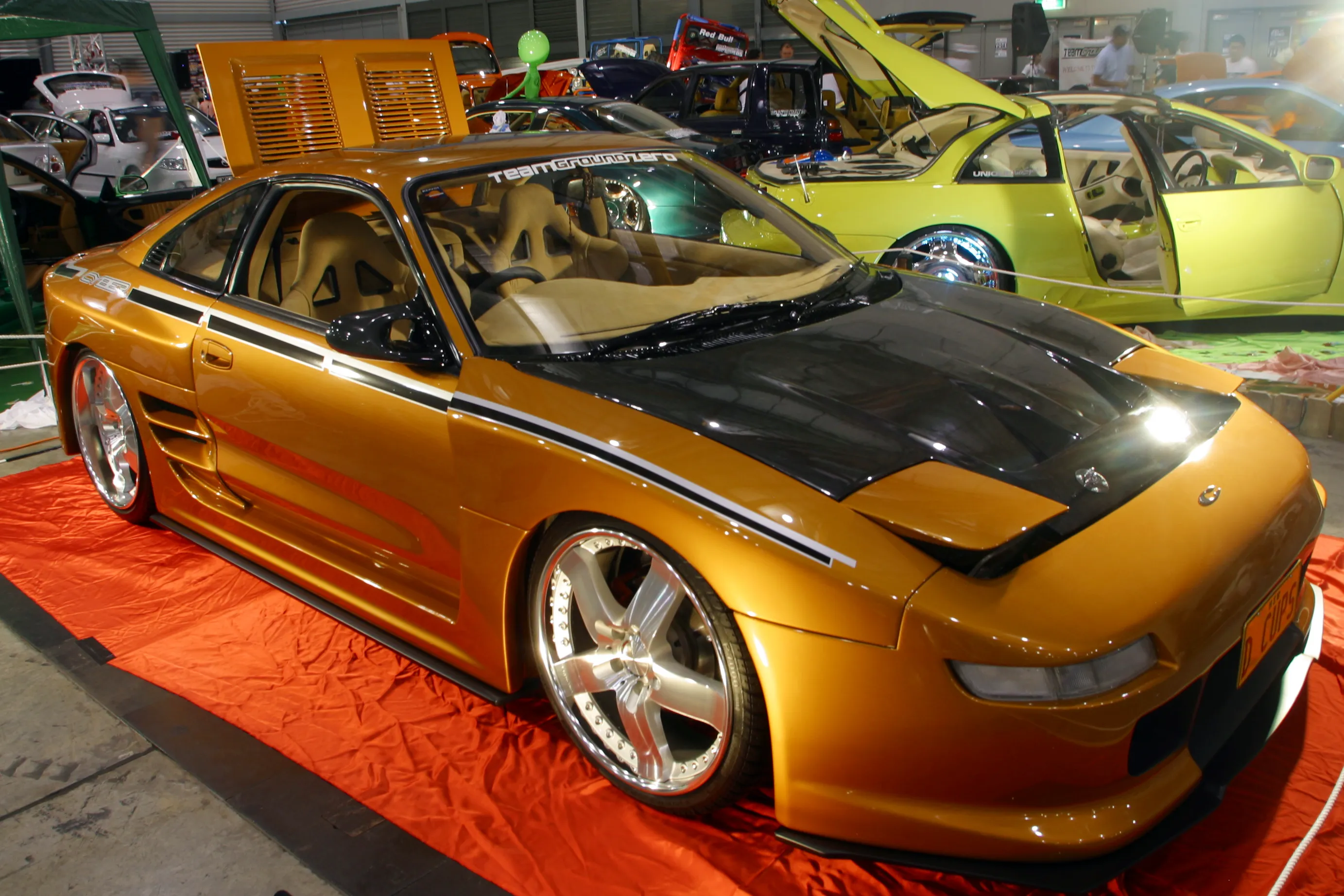 A gold custom MR2 with a black hood and side stripes, large polished alloy wheels, and a "Team Ground Zero" windscreen decal, displayed on an orange sheet at an indoor car show, with other modified cars nearby.