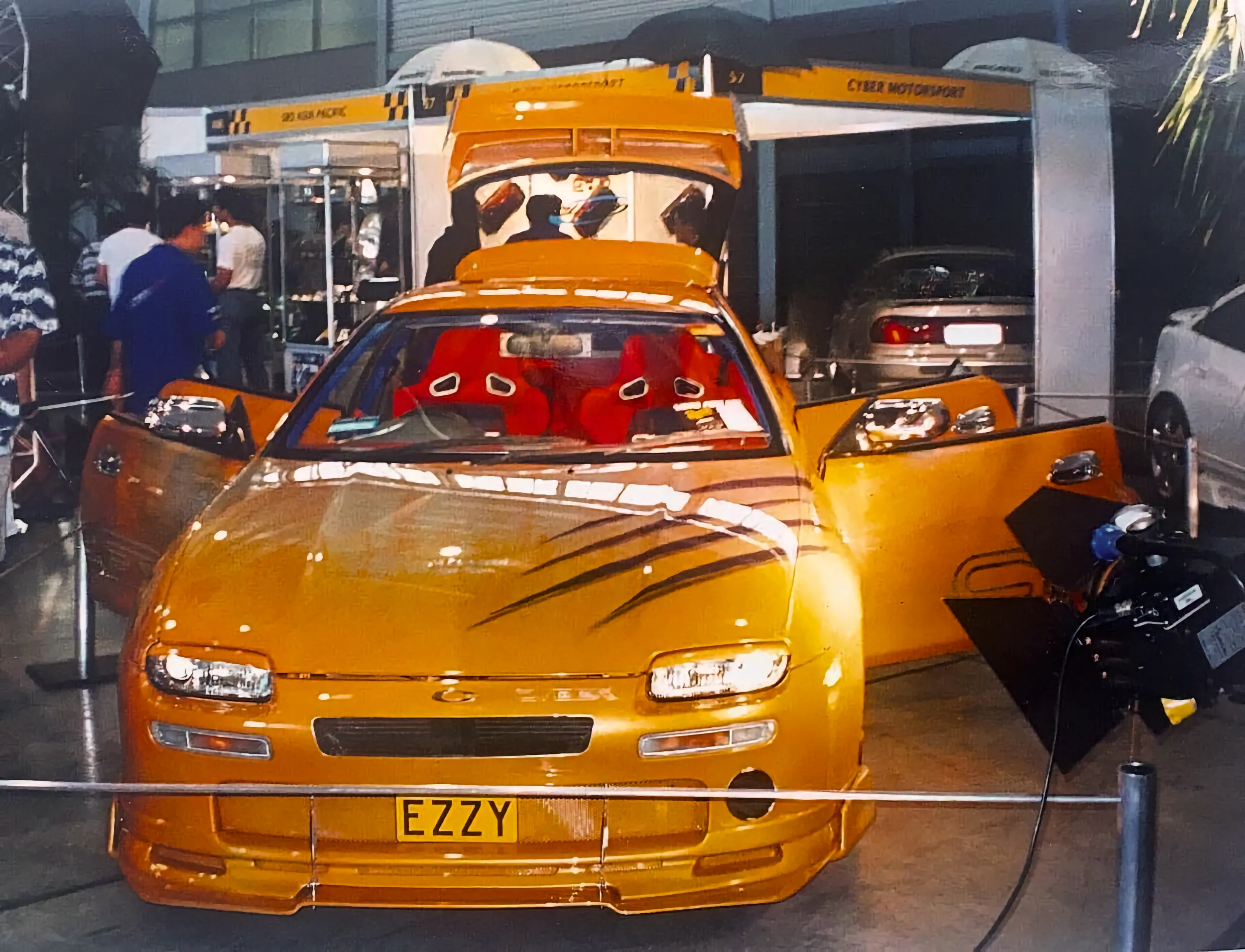 An orange-gold custom sports car displayed with its hood, doors, and rear hatch open, revealing bright red racing seats. It has airbrushed dark stripe graphics on the hood and an "EZZY" yellow number plate, shown at an indoor venue with a "Cyber Motorsport" booth behind it.
