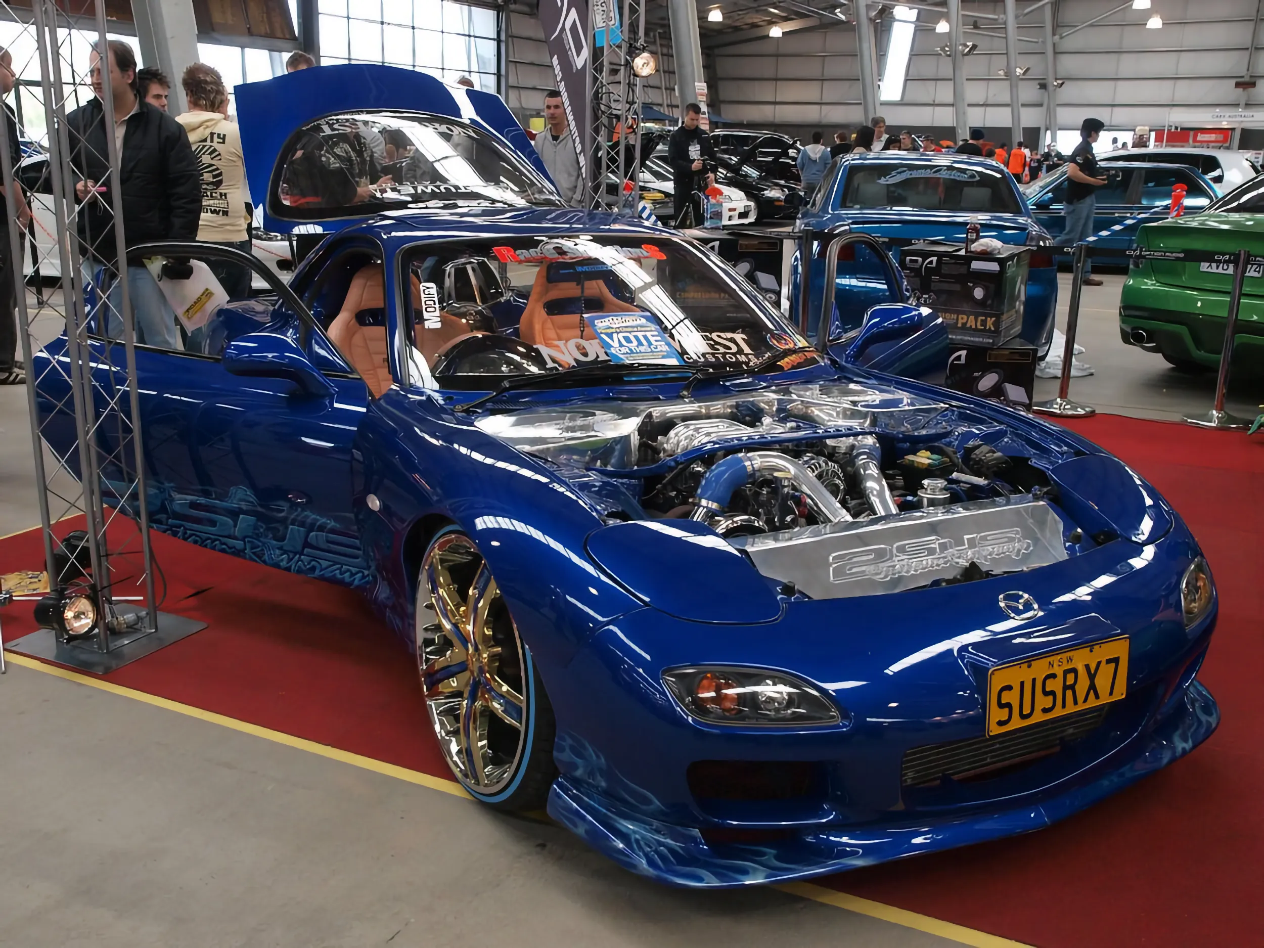 Front three-quarter view of a blue custom sports coupe with its hood open, showing a polished twin-turbo engine bay, tan leather racing seats, and gold-and-chrome wheels. A yellow "SUSRX7" number plate is on the front, and it sits on red carpet at an indoor car show.