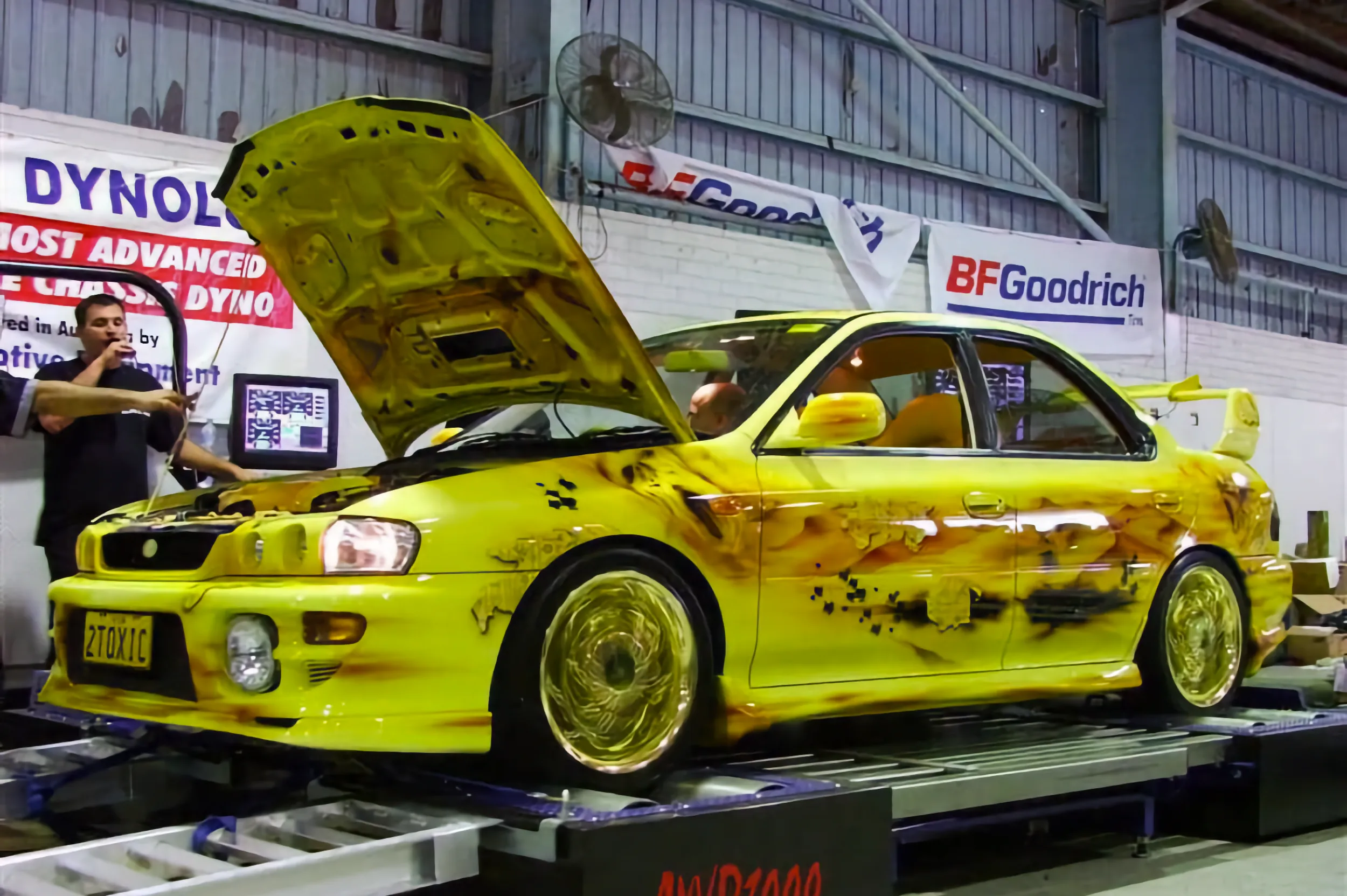 A yellow modified Subaru Impreza with airbrushed flame artwork sits on a chassis dyno, bonnet raised, its "2TOXIC" plate visible, as a technician works nearby under BFGoodrich banners.