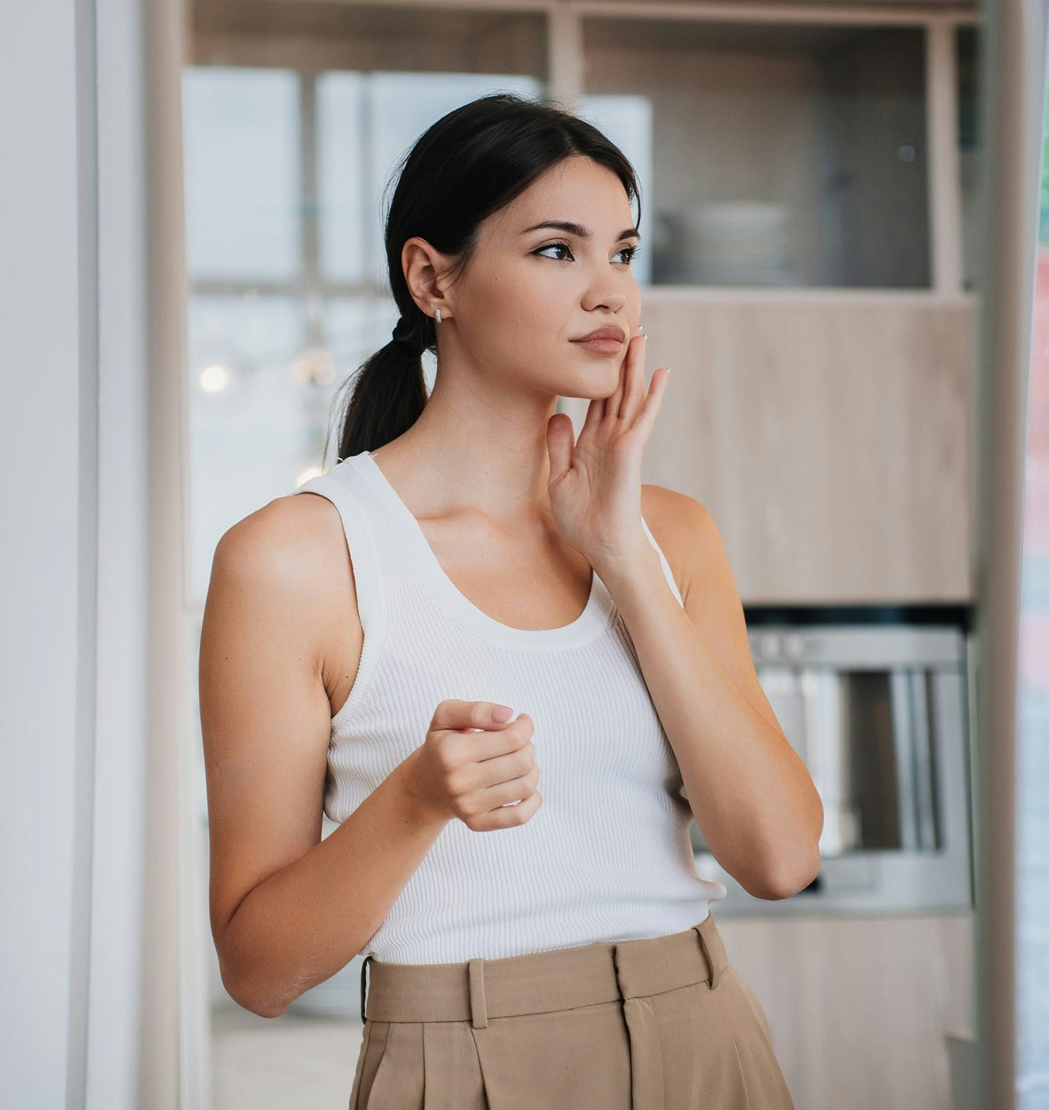 woman in white blouse and chaki pants holds face with left hand while looking left