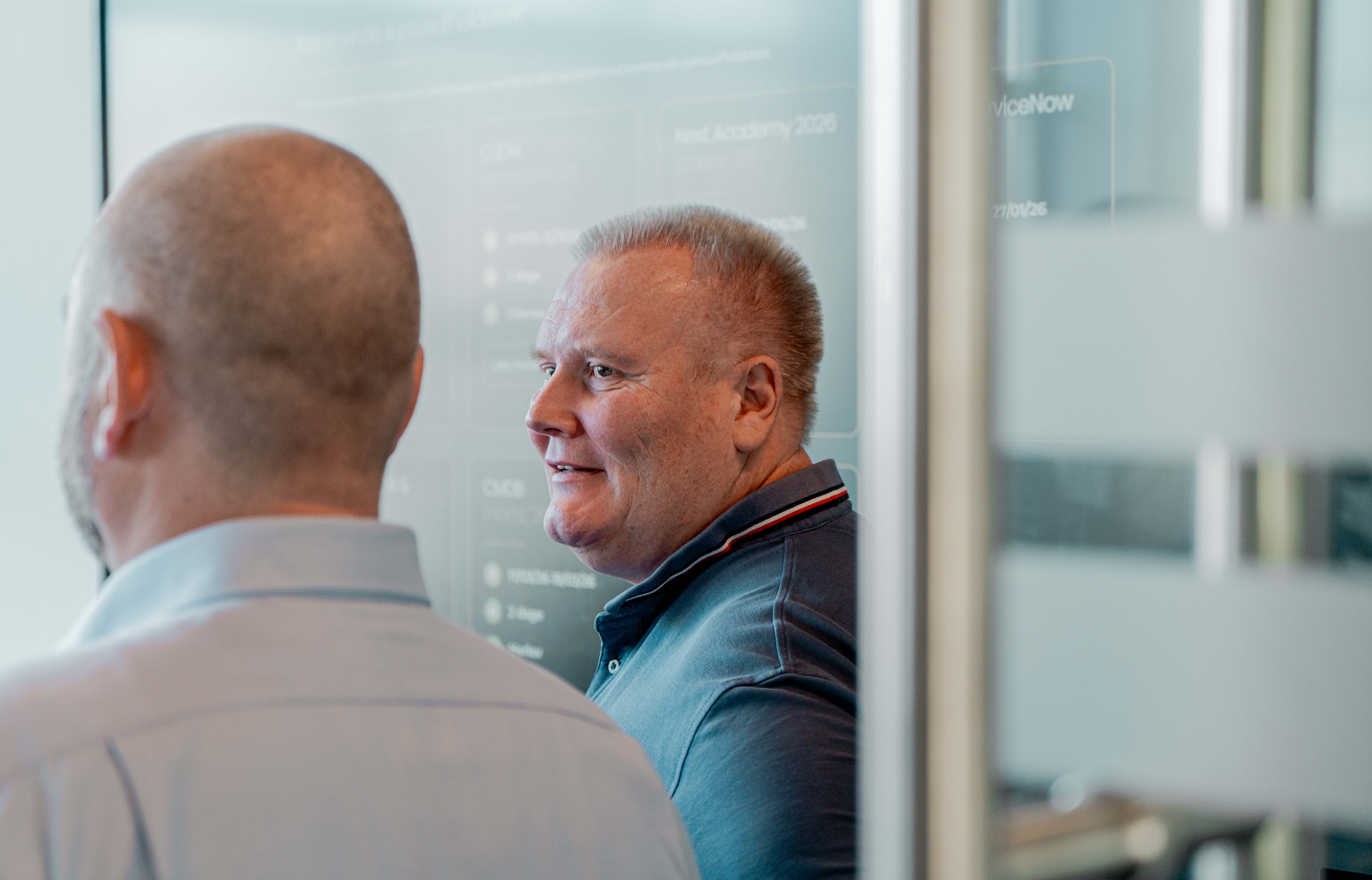 A man in a blue polo shirt smiling in an office with a ServiceNow training screen in the background.