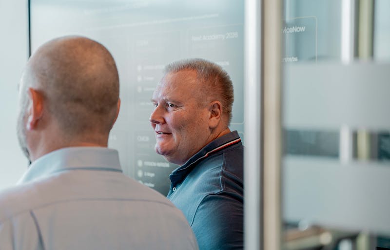A man in a blue polo shirt smiling in an office with a ServiceNow training screen in the background.