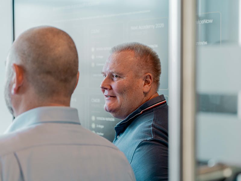 A man in a blue polo shirt smiling in an office with a ServiceNow training screen in the background.