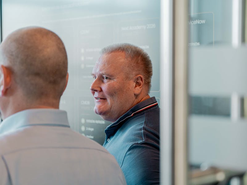 A man in a blue polo shirt smiling in an office with a ServiceNow training screen in the background.