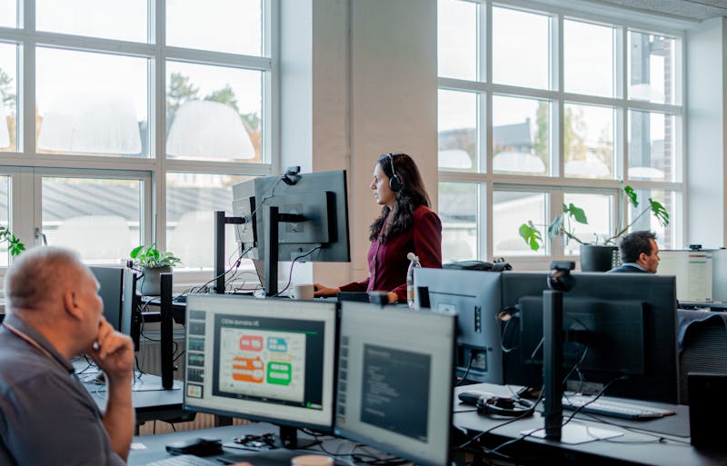 Employees working in an office environment with computer monitors, one of which displays a ServiceNow CSDM domains diagram.
