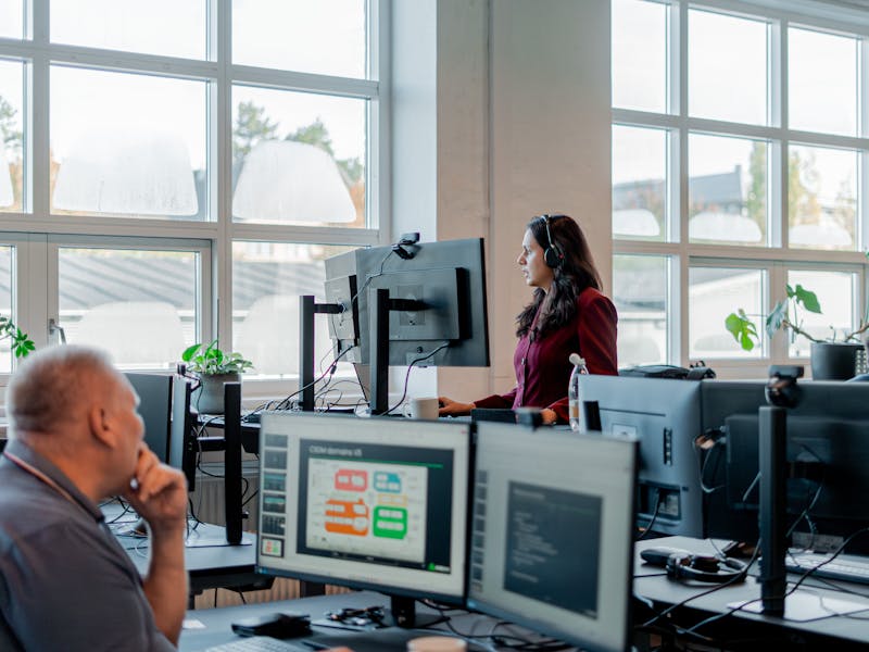 Employees working in an office environment with computer monitors, one of which displays a ServiceNow CSDM domains diagram.