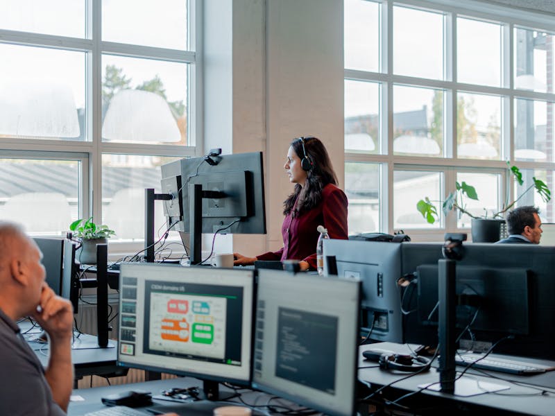 Employees working in an office environment with computer monitors, one of which displays a ServiceNow CSDM domains diagram.