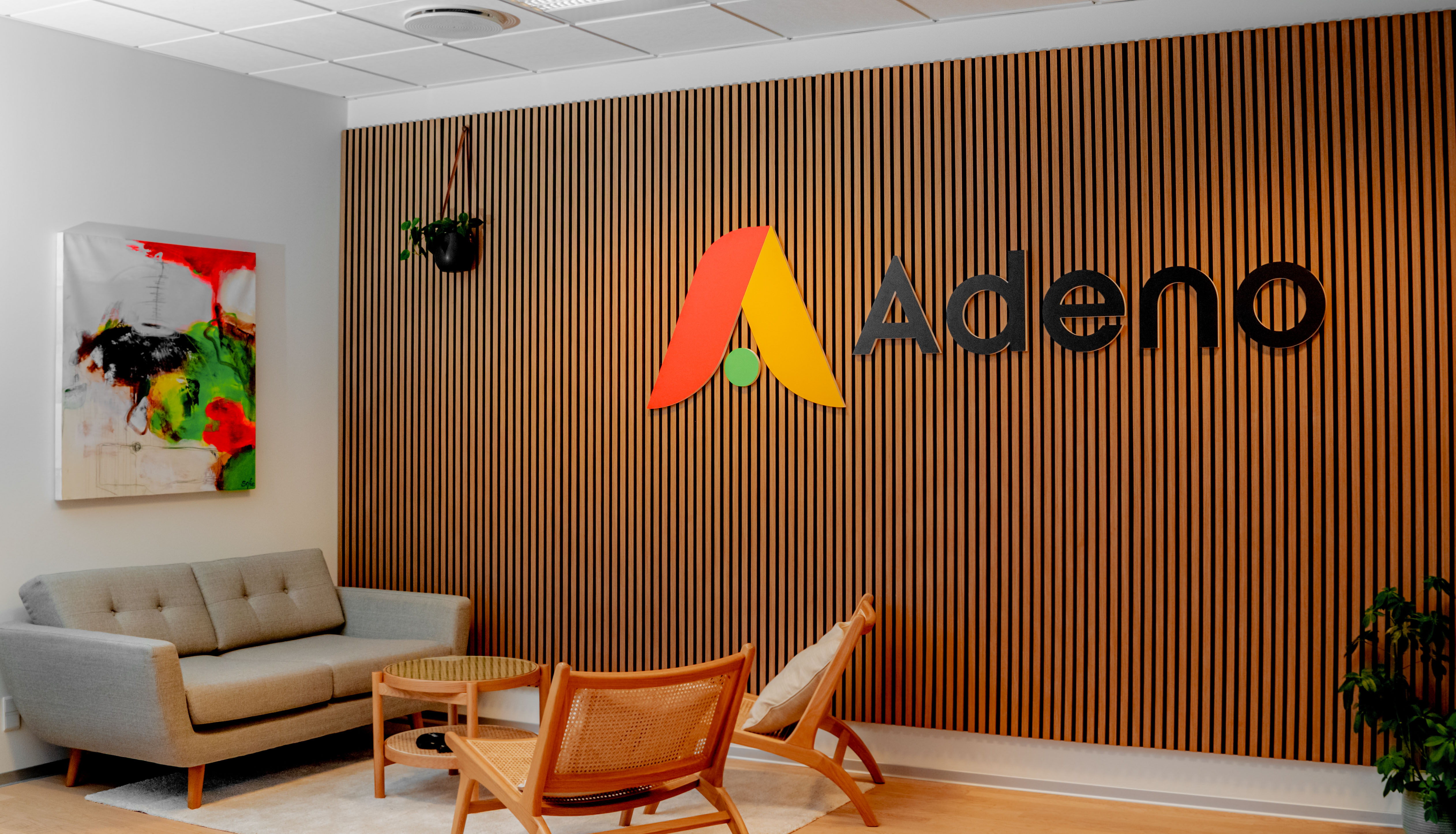 Office interior of Adeno, a ServiceNow partner, featuring a wooden slat wall with the company logo, a grey sofa, and wooden chairs.