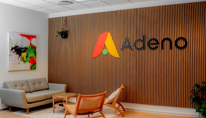 Office interior of Adeno, a ServiceNow partner, featuring a wooden slat wall with the company logo, a grey sofa, and wooden chairs.