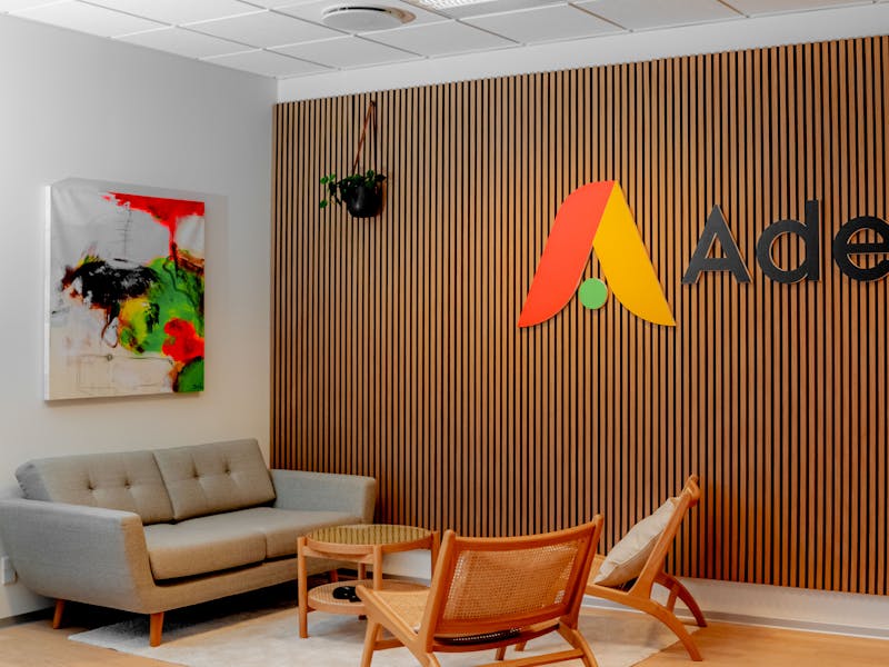 Office interior of Adeno, a ServiceNow partner, featuring a wooden slat wall with the company logo, a grey sofa, and wooden chairs.