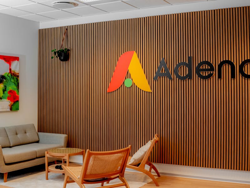 Office interior of Adeno, a ServiceNow partner, featuring a wooden slat wall with the company logo, a grey sofa, and wooden chairs.