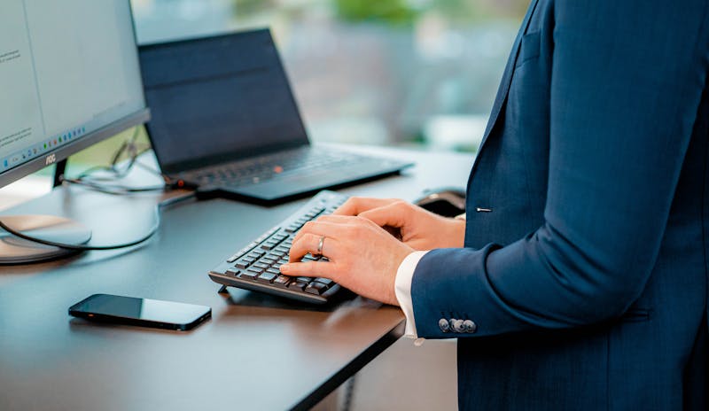 A person in a blue suit typing on a keyboard at a desk with a monitor and laptop, representing a ServiceNow professional.