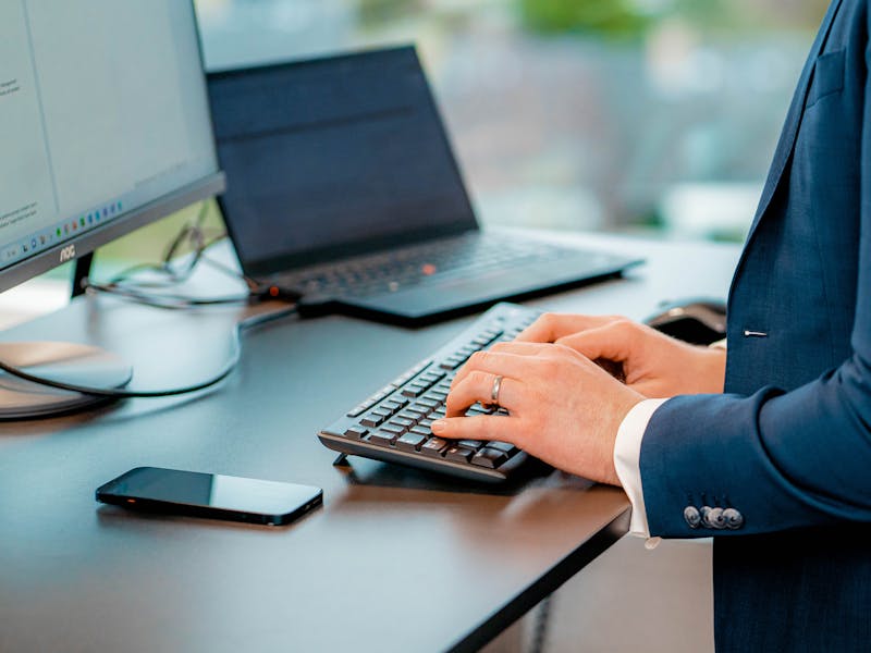 A person in a blue suit typing on a keyboard at a desk with a monitor and laptop, representing a ServiceNow professional.