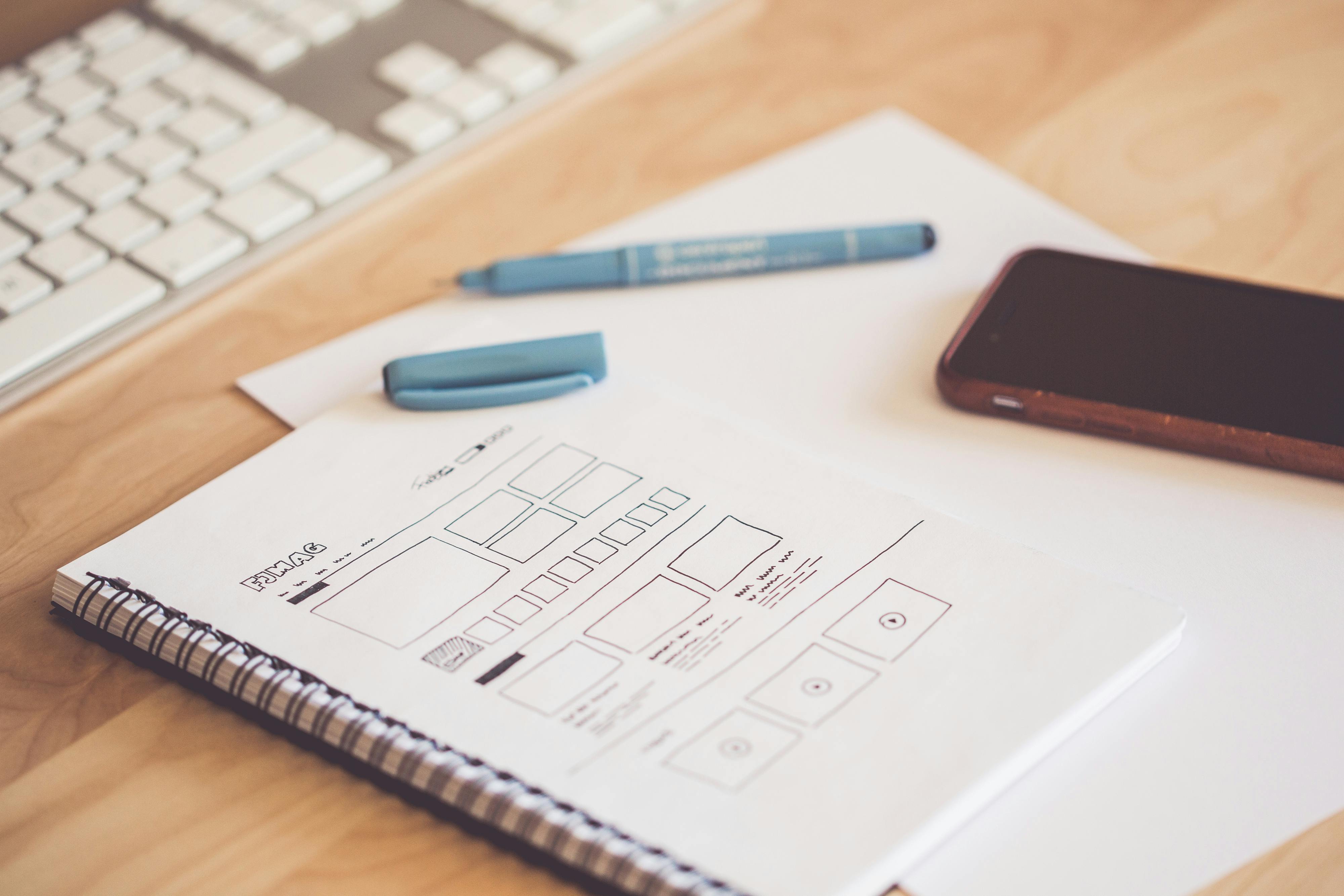 A desk with a keyboard, smartphone, and a notebook showing a hand-drawn ServiceNow UI wireframe.