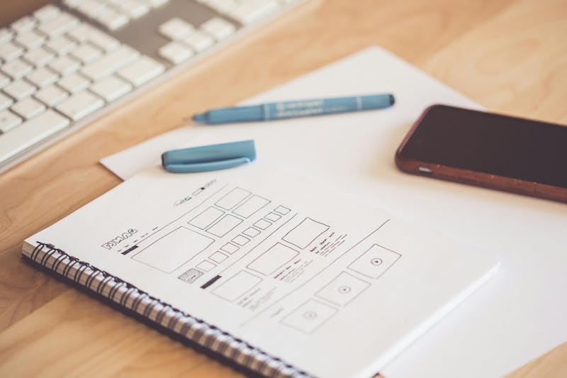 A desk with a keyboard, smartphone, and a notebook showing a hand-drawn ServiceNow UI wireframe.