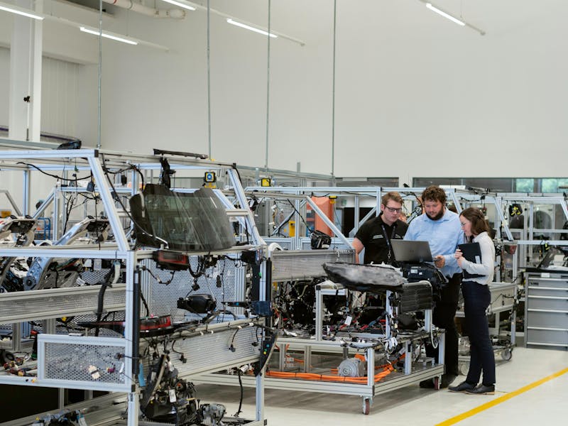 Three people in an industrial facility using a laptop near automotive testing rigs, illustrating ServiceNow.