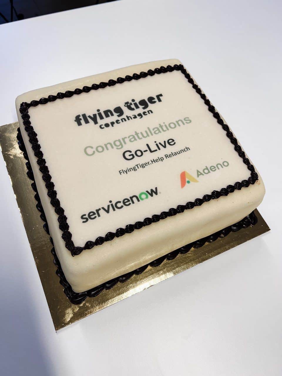 A square celebration cake with white frosting featuring the Flying Tiger Copenhagen, ServiceNow, and Adeno logos and text celebrating the relaunch of FlyingTiger.Help.