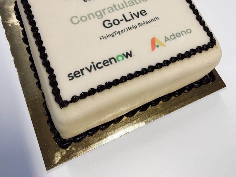 A square celebration cake with white frosting featuring the Flying Tiger Copenhagen, ServiceNow, and Adeno logos and text celebrating the relaunch of FlyingTiger.Help.