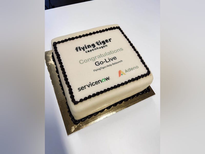 A square celebration cake with white frosting featuring the Flying Tiger Copenhagen, ServiceNow, and Adeno logos and text celebrating the relaunch of FlyingTiger.Help.