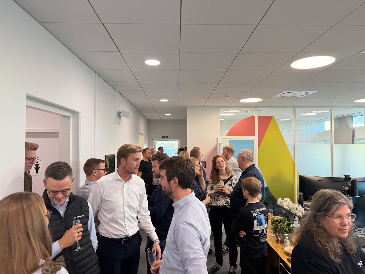 People socializing in a modern office during a housewarming event, with a ServiceNow logo visible on a person's vest.