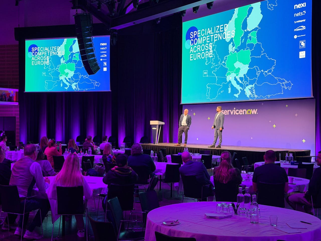 Two speakers on a stage at a ServiceNow event with a screen displaying a map of Europe and an audience seated at tables.