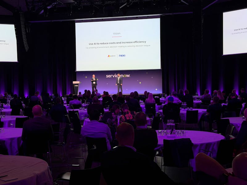 Two speakers on a stage presenting a slide about AI to an audience at a ServiceNow event.