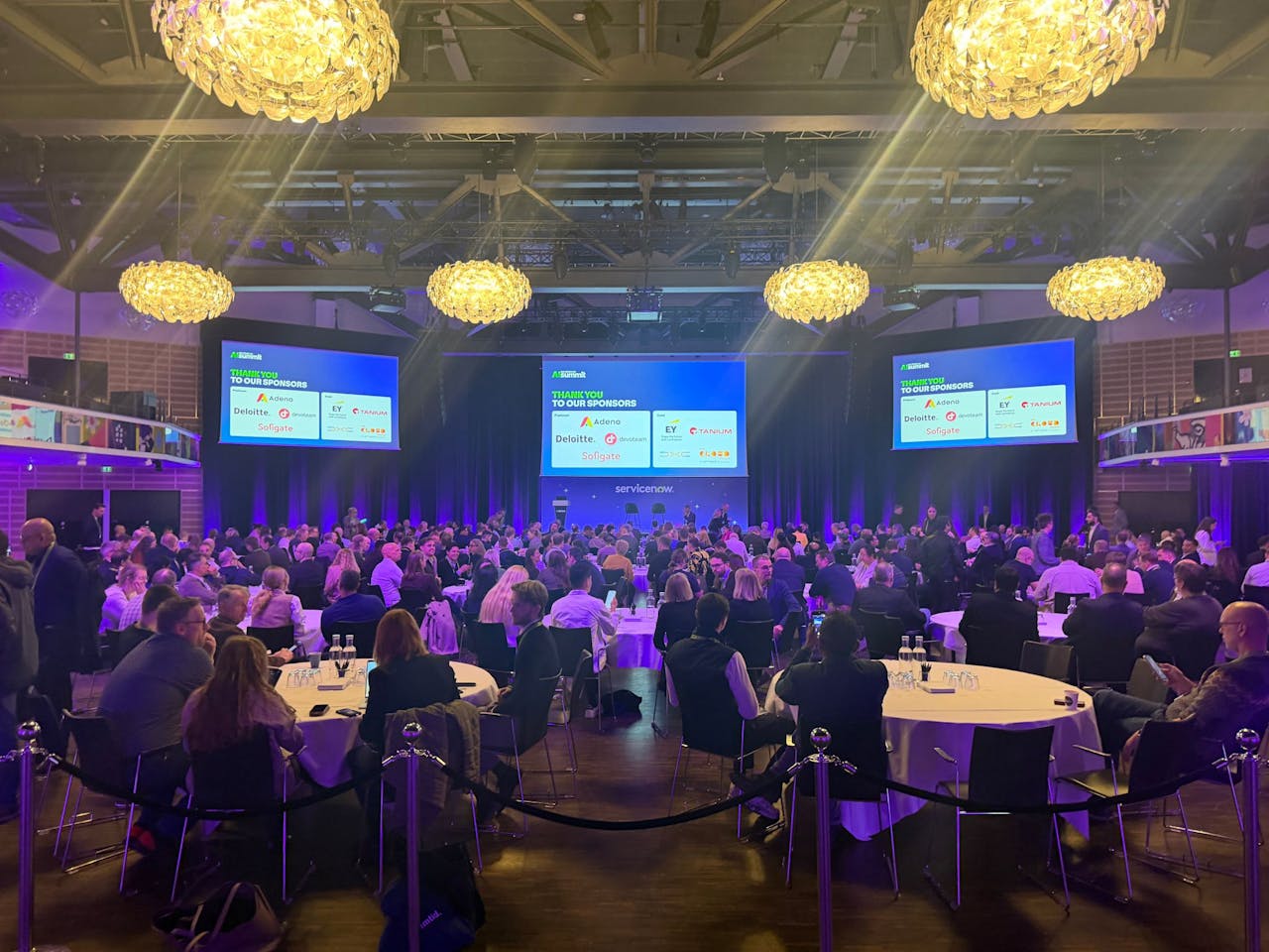 An audience seated at round tables in a conference hall during a ServiceNow summit.
