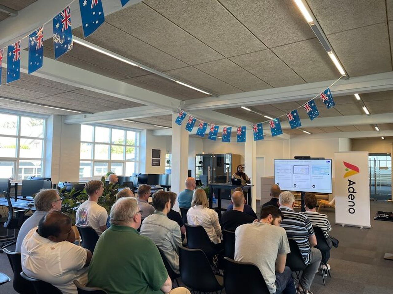 People attending a ServiceNow Australia Release Event presentation in an office with Australian flags and an Adeno banner.