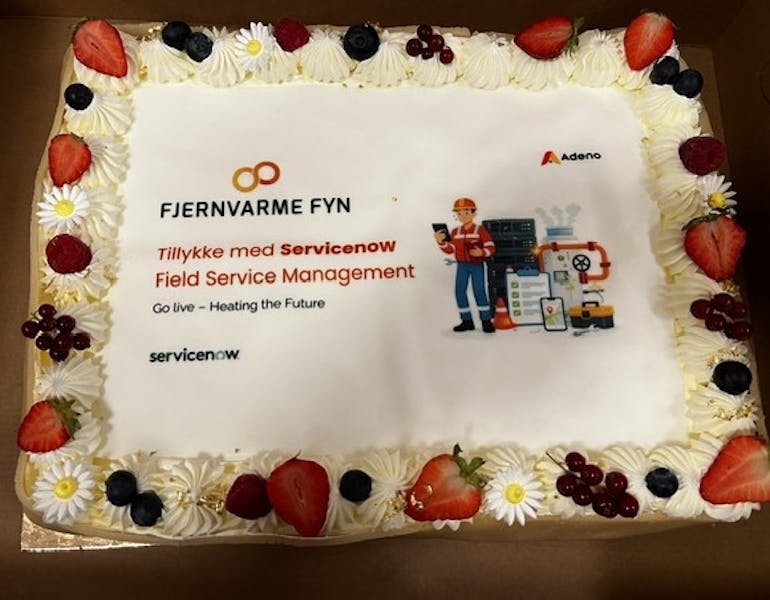 A celebratory cake decorated with fruit and flowers for the Fjernvarme Fyn and ServiceNow Field Service Management go-live, featuring the Adeno logo.