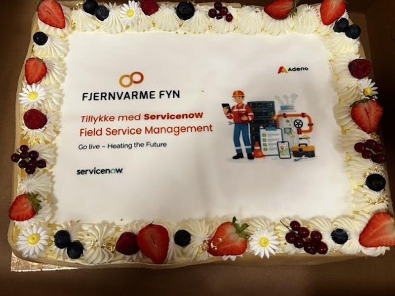 A celebratory cake decorated with fruit and flowers for the Fjernvarme Fyn and ServiceNow Field Service Management go-live, featuring the Adeno logo.