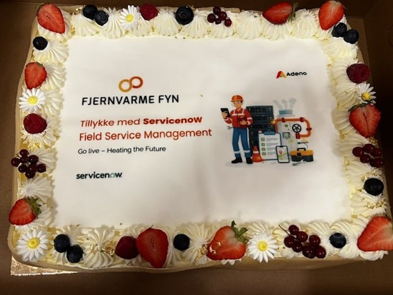 A celebratory cake decorated with fruit and flowers for the Fjernvarme Fyn and ServiceNow Field Service Management go-live, featuring the Adeno logo.