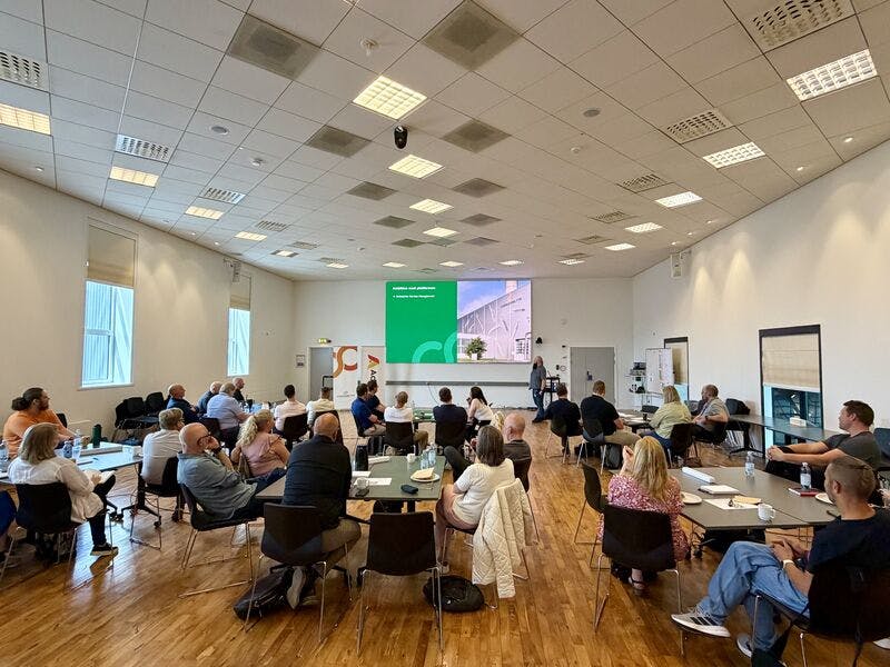 A group of people attending a ServiceNow PO Network meeting at Fjernvarme Fyn, seated at tables and watching a presentation on a large screen.