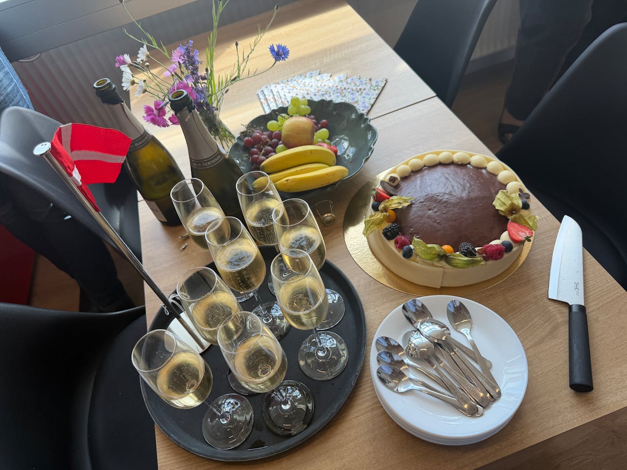 A celebratory spread on a wooden table for a ServiceNow event, featuring champagne, a chocolate cake, fruit, and a Danish flag.