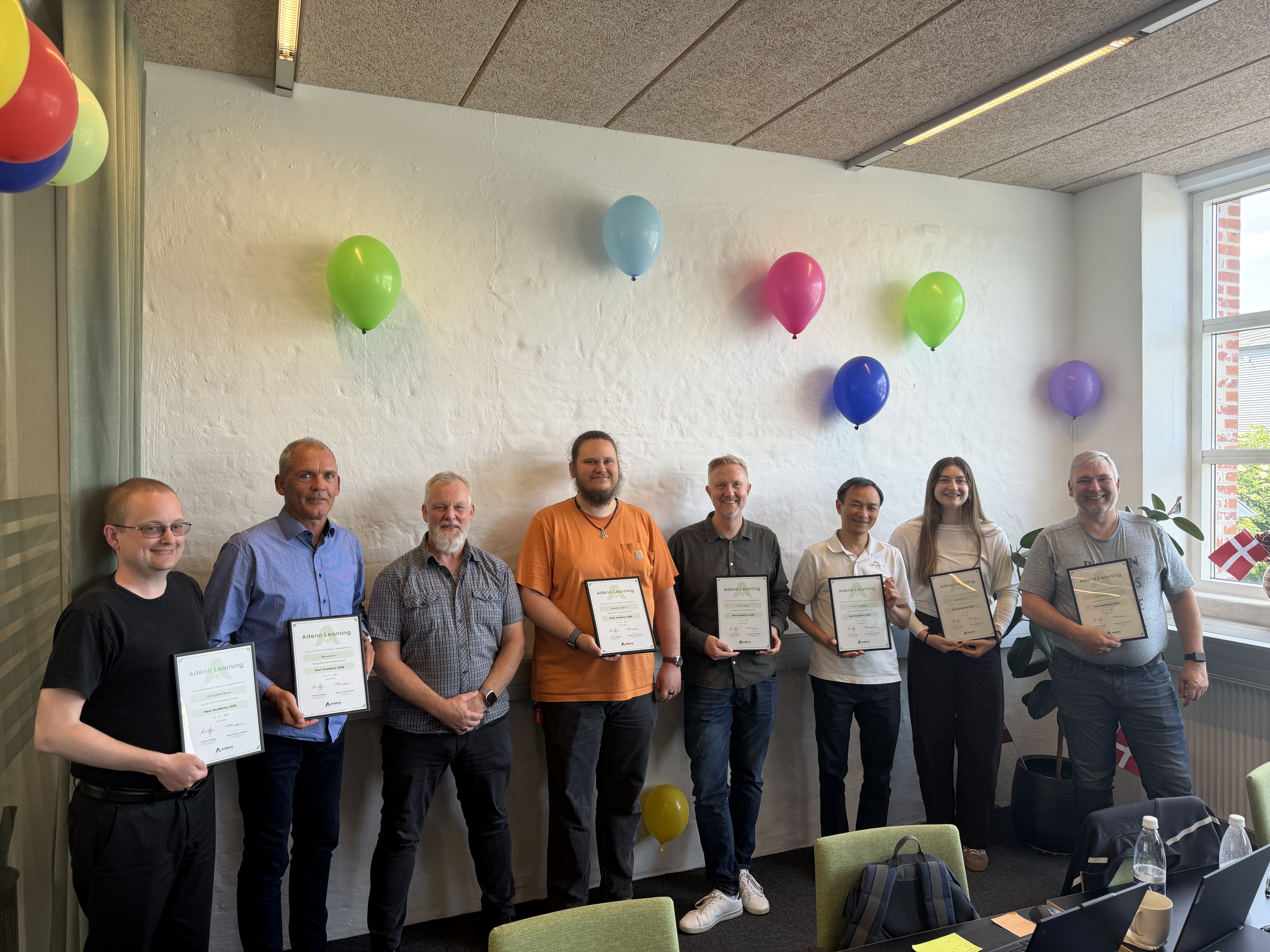 Eight people holding certificates at the Next Academy 2026 graduation for ServiceNow Partner Adeno in a room with balloons."