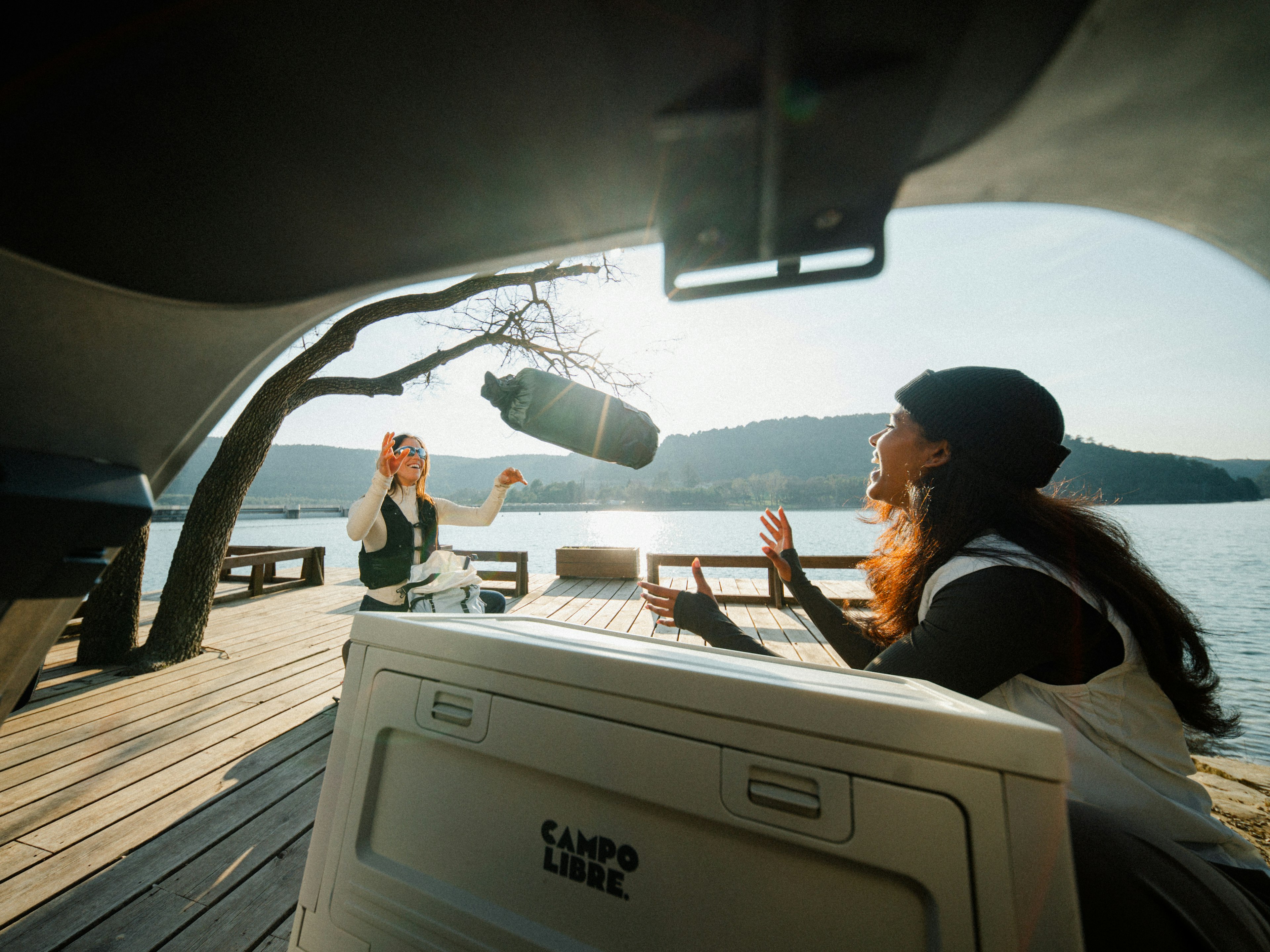 Two people conversing on a shaded deck overlooking a calm lake, captured from behind a large object.