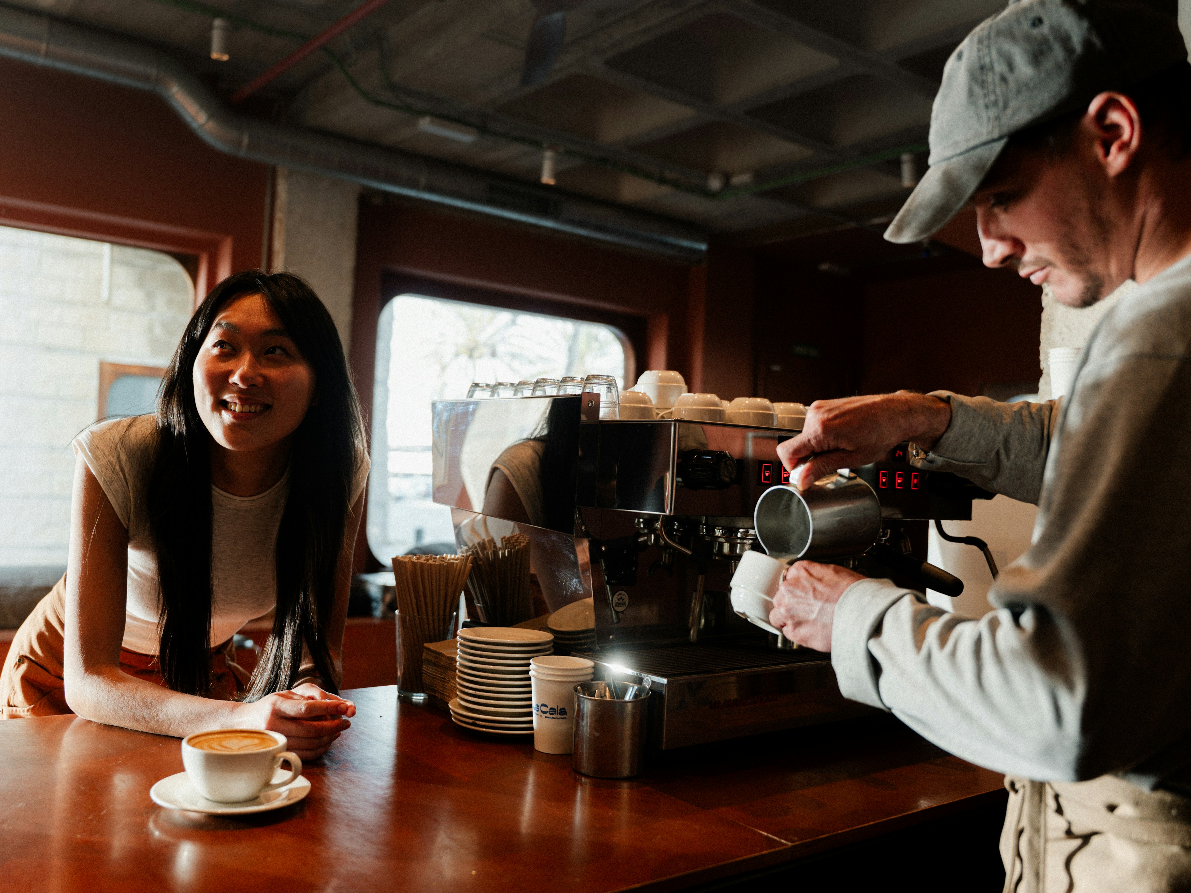 Un barista prepara il caffè mentre una donna sorride al bancone in un'accogliente caffetteria.