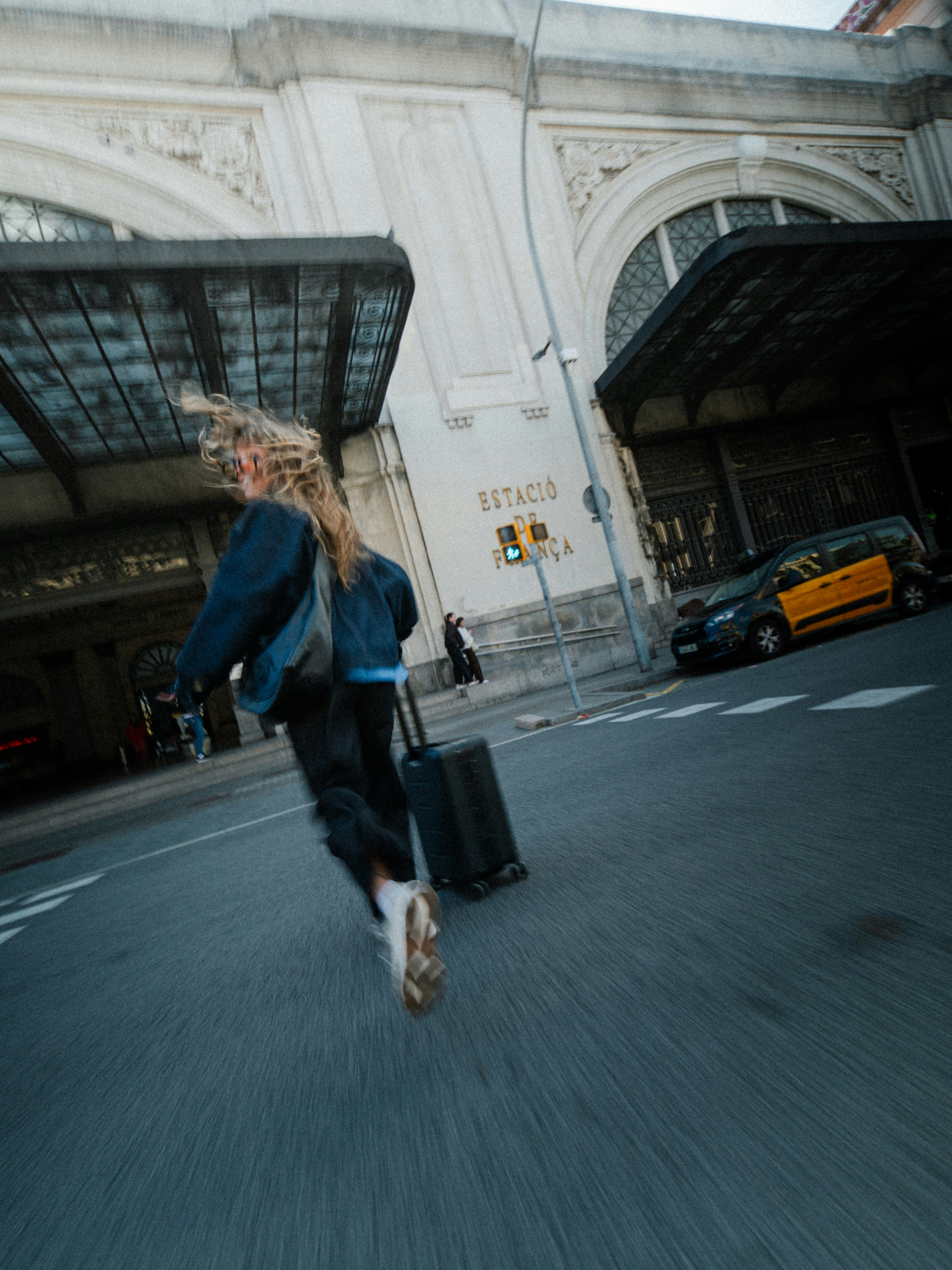 Una persona con una maleta camina hacia un edificio grande y ornamentado con ventanas en arco y un taxi aparcado cerca.